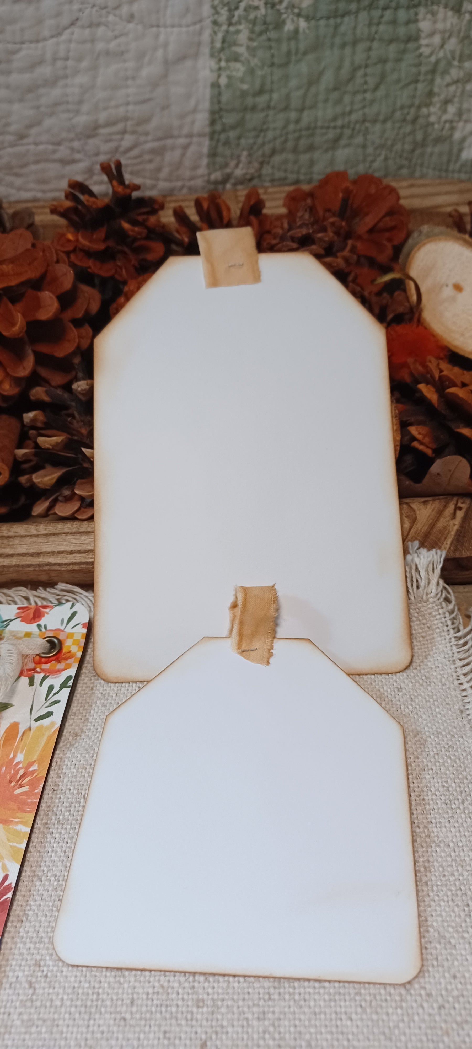 Two blank white wooden tag-shaped photo frames with natural wood edges and small tan tabs at the top, displayed on a burlap surface with autumn decor in the background.