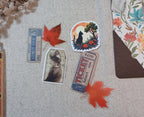 A styled flat-lay of vintage-style paper cutouts and stickers on a beige linen fabric background, featuring two blue admission tickets, a black cat silhouette sticker under a tree, a sepia-toned ticket, and orange autumn leaves.