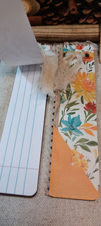 A close-up of a handmade bookmark or card made from patterned floral paper, burlap, and orange cardstock, shown next to a lined paper strip on a woven fabric background.