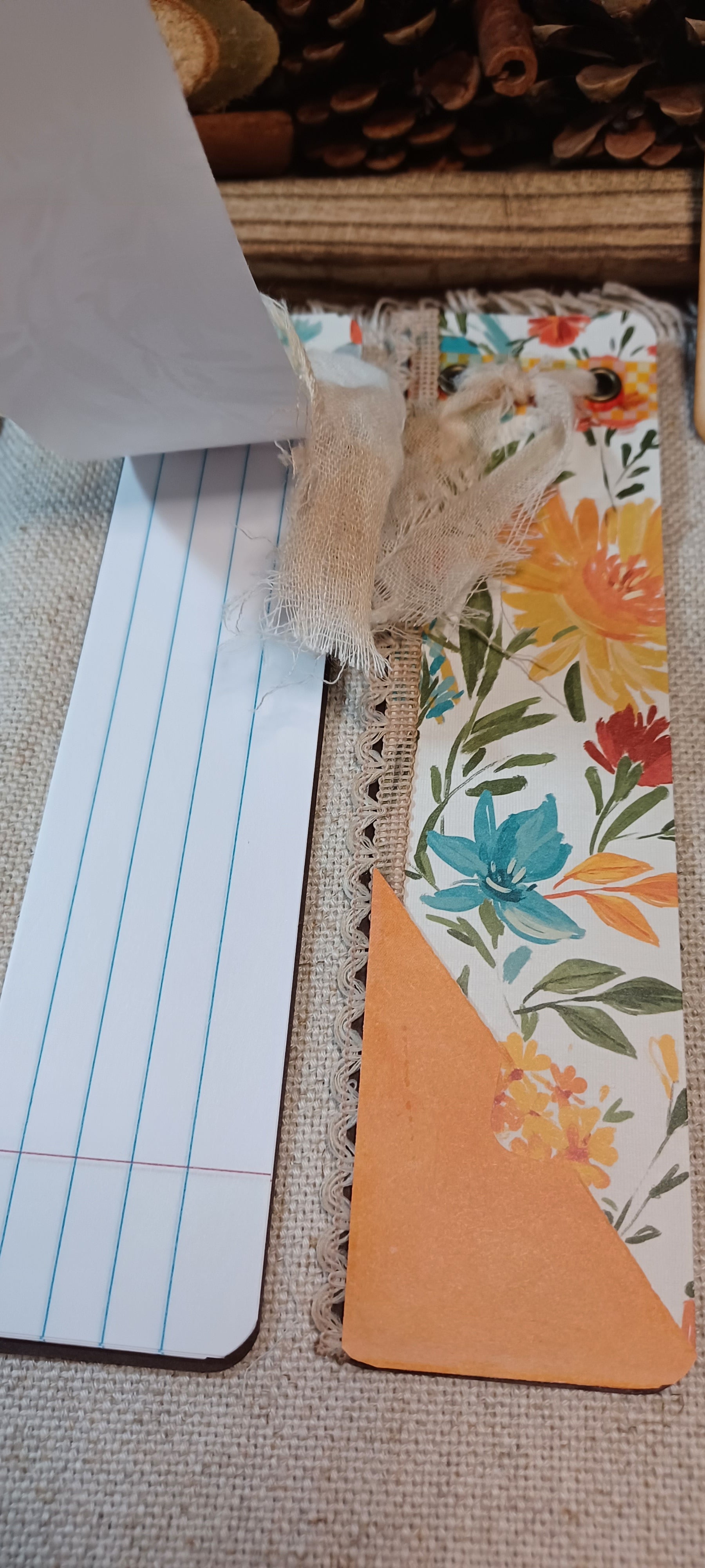 A close-up of a handmade bookmark or card made from patterned floral paper, burlap, and orange cardstock, shown next to a lined paper strip on a woven fabric background.