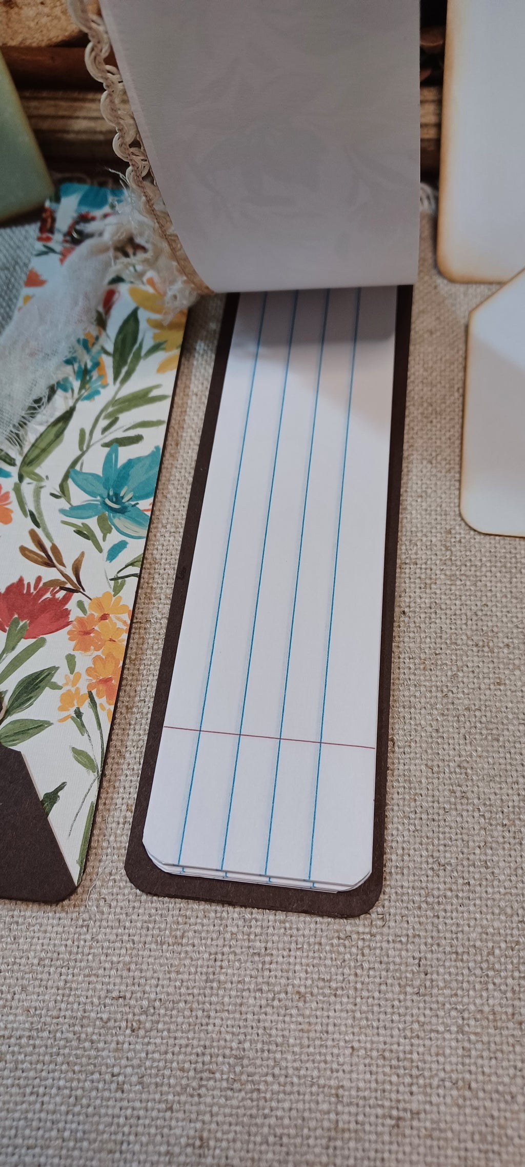 A close-up of a rectangular lined notepad or planner insert with blue and red lines, placed on a beige woven surface next to floral-patterned paper and other partial objects.