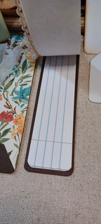 A close-up of a rectangular lined notepad or planner insert with blue and red lines, placed on a beige woven surface next to floral-patterned paper and other partial objects.