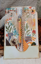 A decorative handmade journal or scrapbook page with floral paper, burlap ribbon, vintage-style stickers, and a matching floral pen placed in the center, photographed on a fabric surface with rustic props around it.