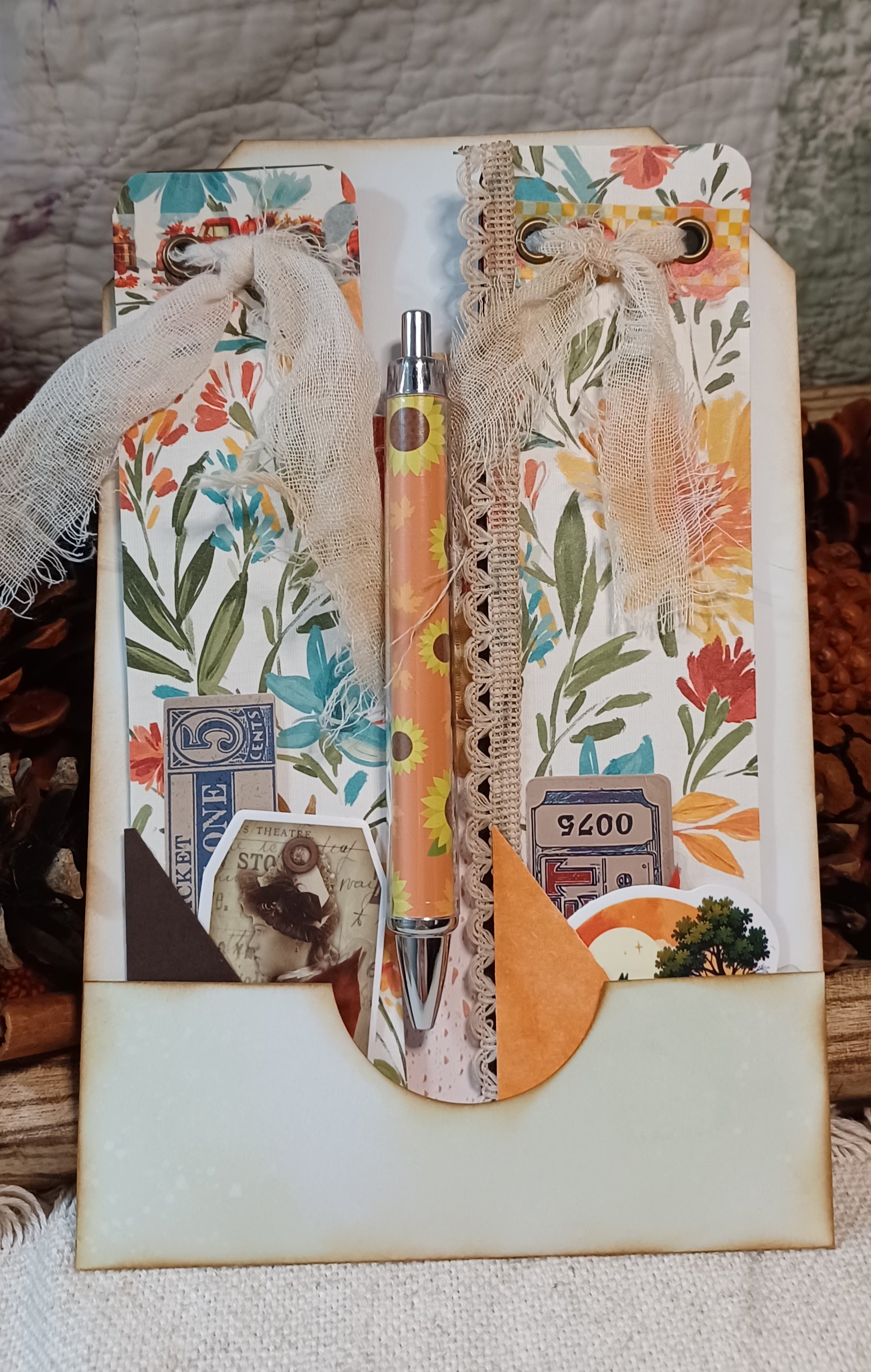 A decorative handmade journal or scrapbook page with floral paper, burlap ribbon, vintage-style stickers, and a matching floral pen placed in the center, photographed on a fabric surface with rustic props around it.