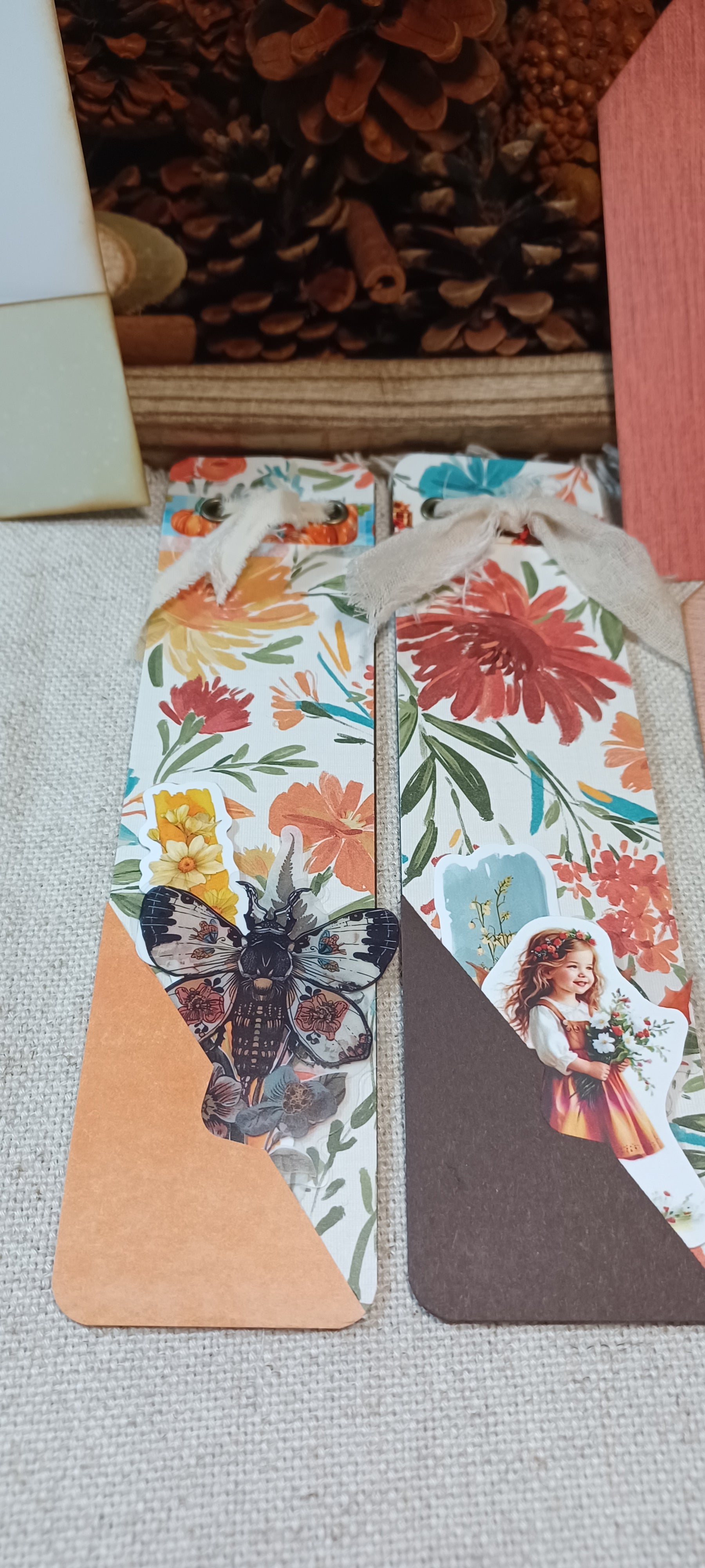 Two decorative paper bookmarks with floral prints, butterflies, and vintage-style illustrations, laid flat on a textured fabric surface with pinecones in the background.