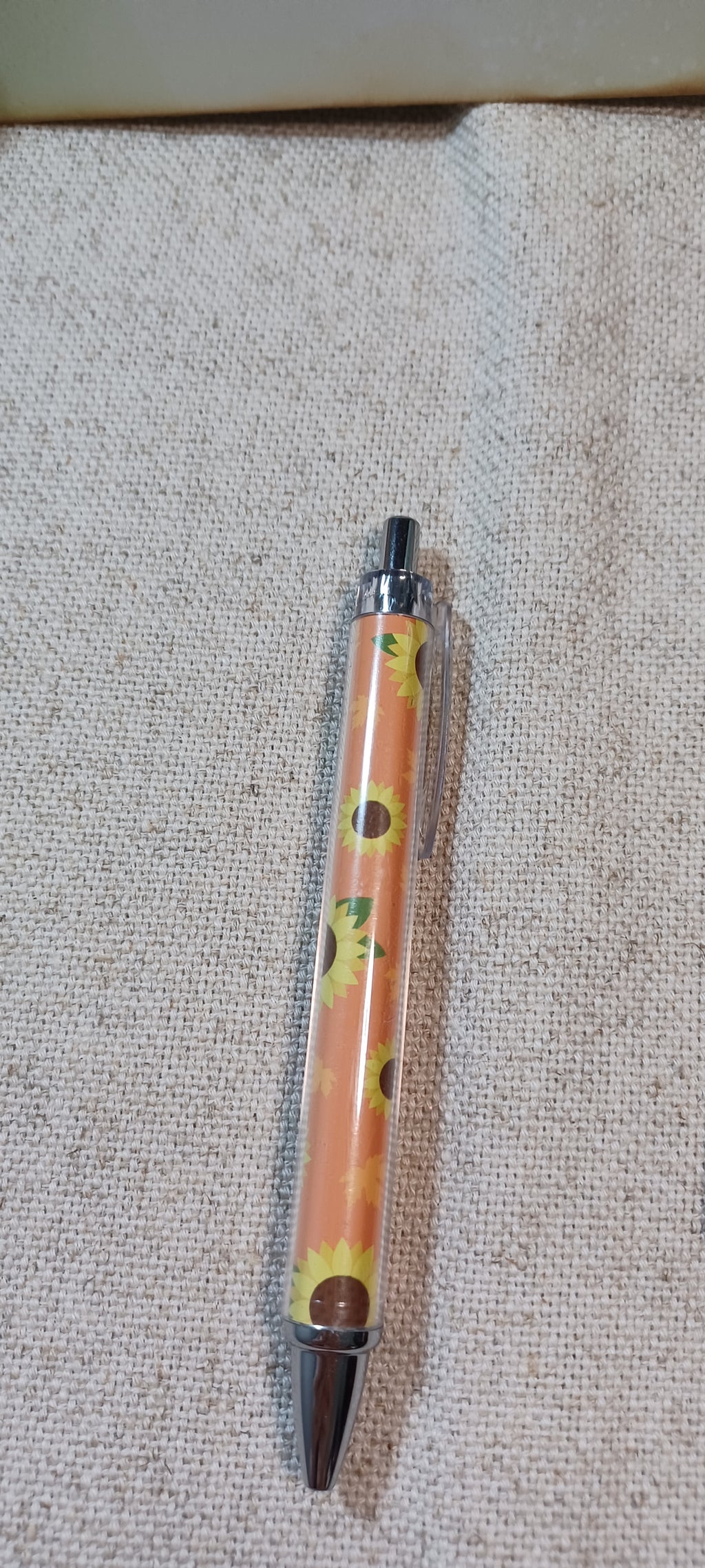 A retractable pen with a clear barrel showing a yellow floral pattern  on burnt umber, capped with silver metal and a dark brown tip, lying on a textured beige fabric background.