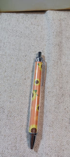 A retractable pen with a clear barrel showing a yellow floral pattern  on burnt umber, capped with silver metal and a dark brown tip, lying on a textured beige fabric background.