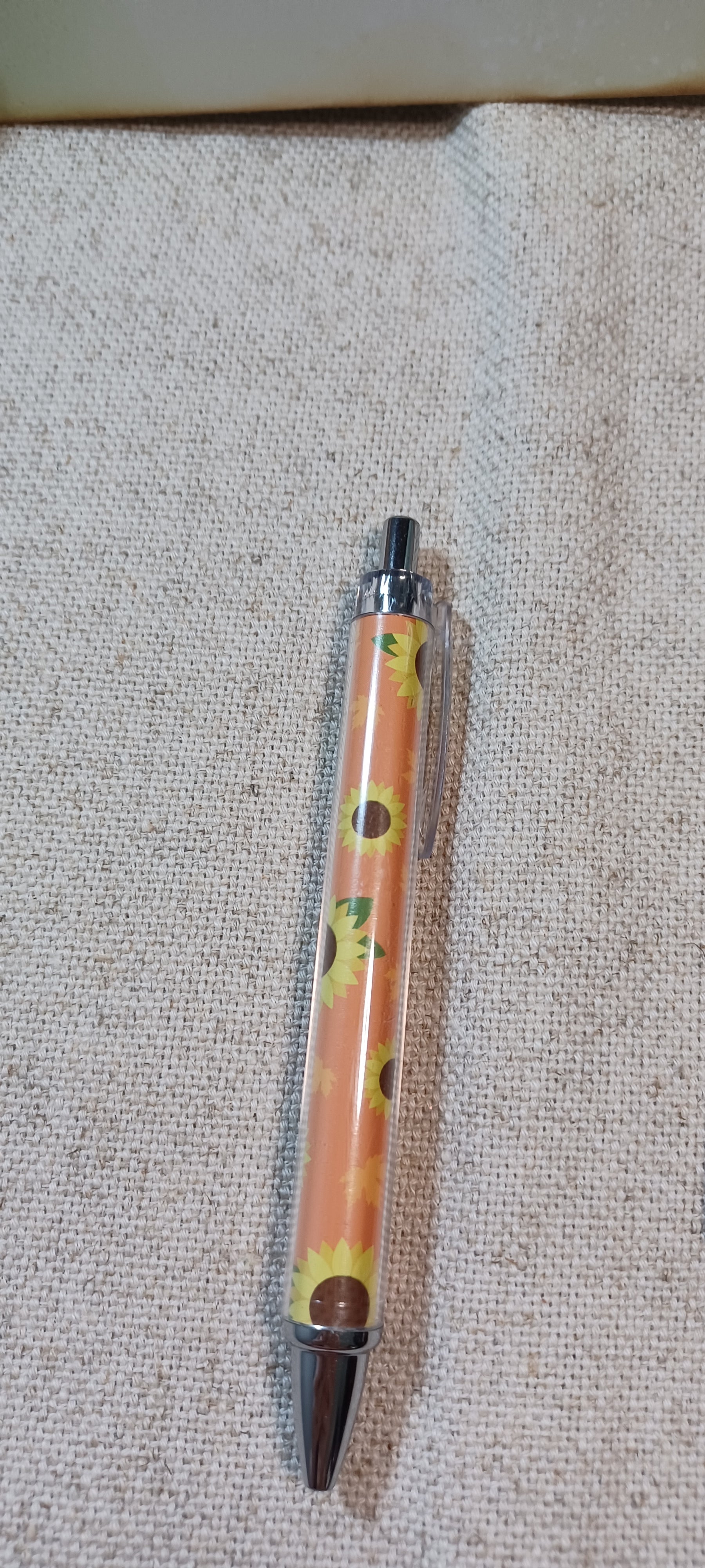 A retractable pen with a clear barrel showing a yellow floral pattern  on burnt umber, capped with silver metal and a dark brown tip, lying on a textured beige fabric background.
