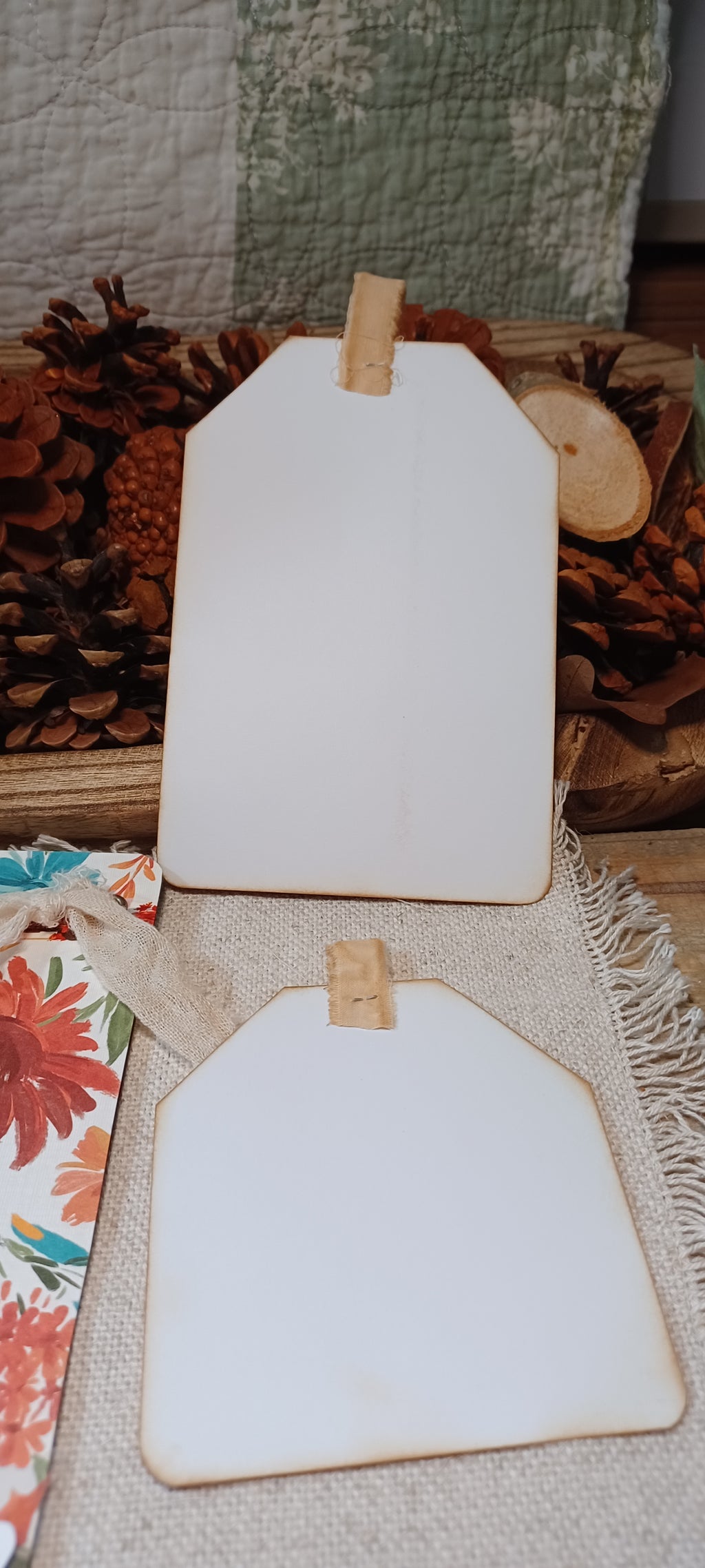 Two blank white wooden gift tags with natural wood edges and small wooden clips, displayed on a burlap surface with pinecones and floral paper in the background.