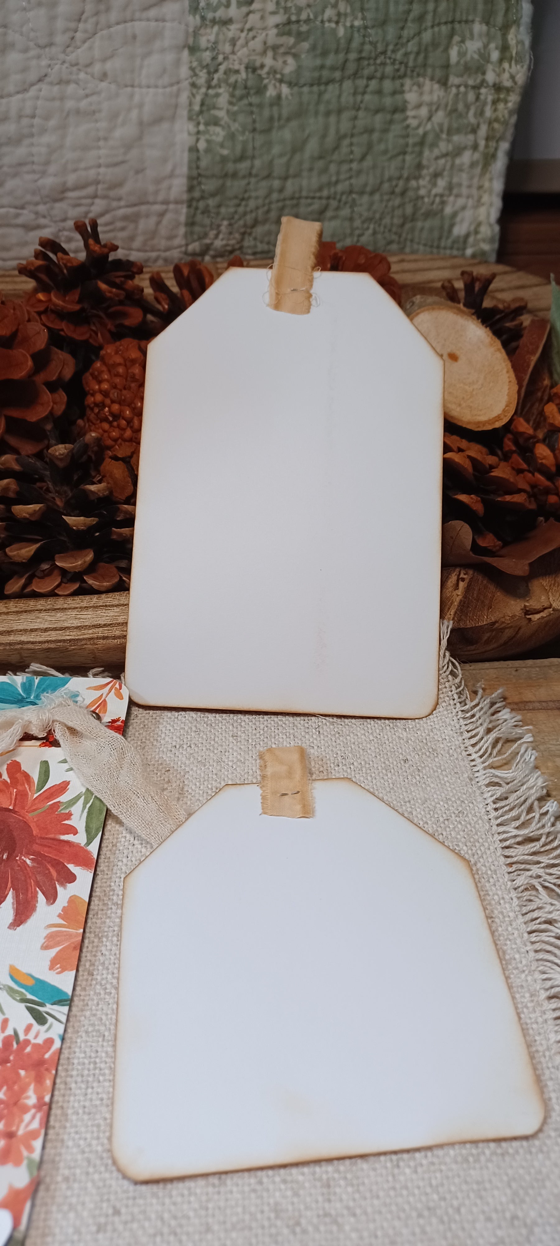 Two blank white wooden gift tags with natural wood edges and small wooden clips, displayed on a burlap surface with pinecones and floral paper in the background.