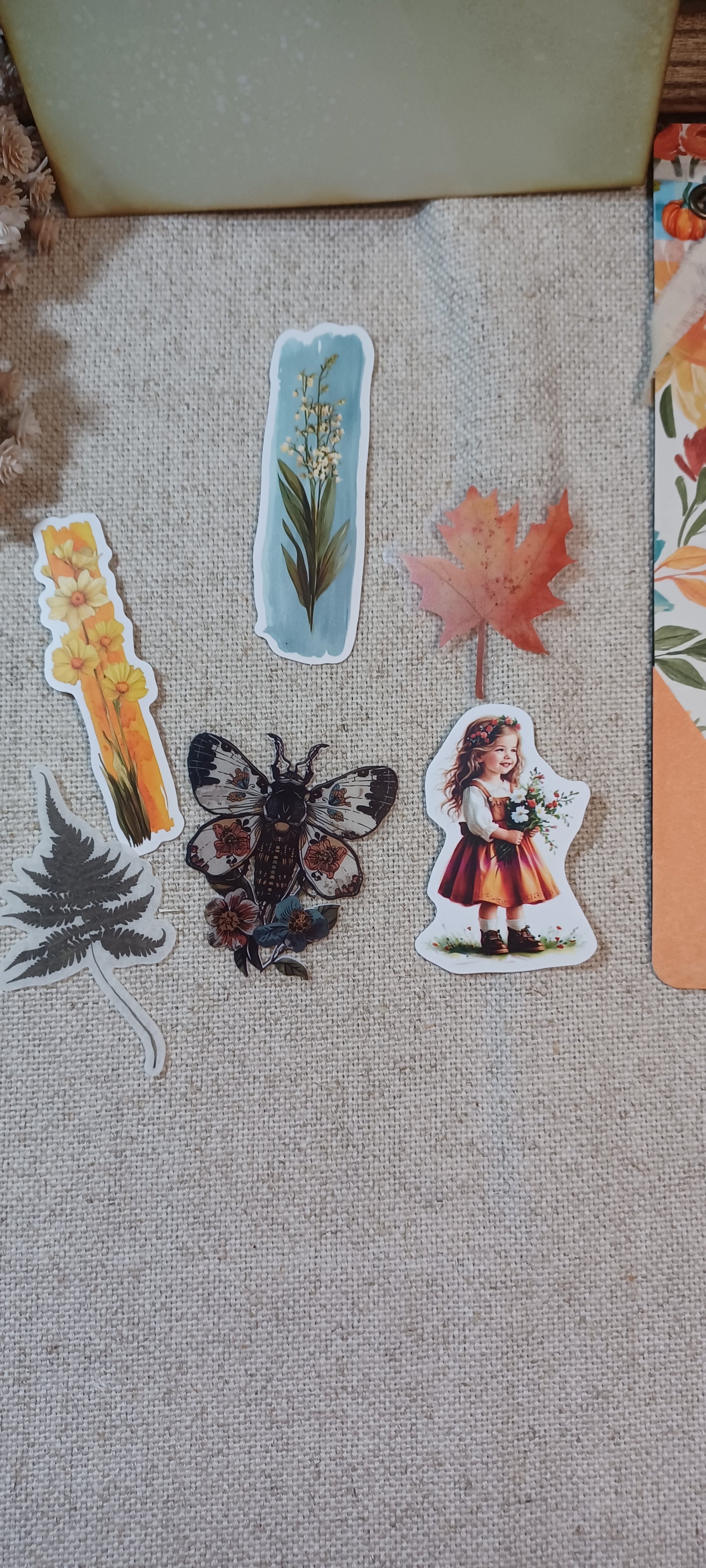 A set of autumn-themed die-cut stickers arranged on a beige linen fabric background. The stickers include a yellow autumn leaf, a black and brown butterfly, a gray pine tree, a light blue flower stem, a red maple leaf, and a vintage-style illustration of a girl in a red dress.