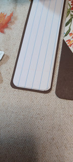 A close-up of a rectangular lined notepad or bookmark with rounded corners, placed on a textured beige fabric surface next to other paper items.