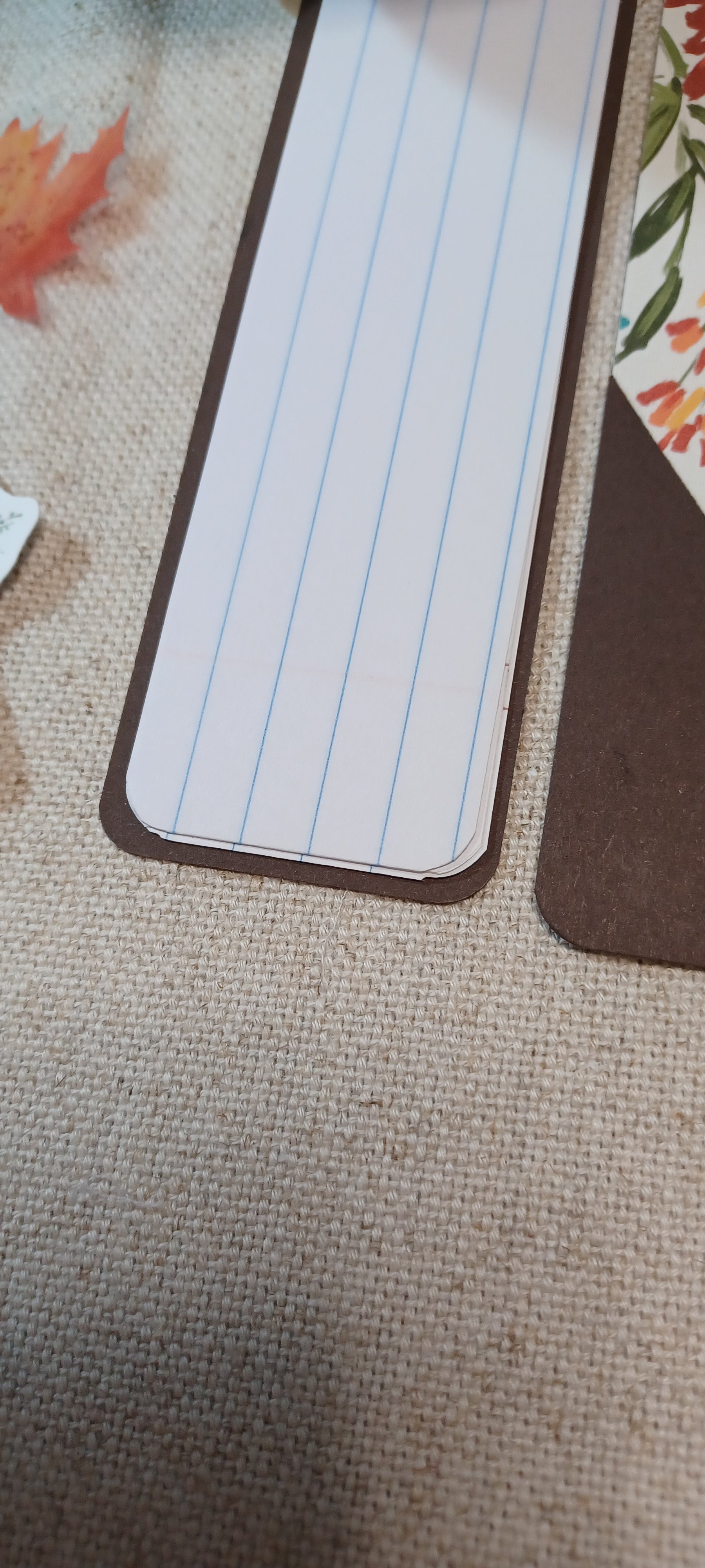A close-up of a rectangular lined notepad or bookmark with rounded corners, placed on a textured beige fabric surface next to other paper items.