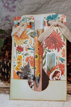A decorative gift set featuring a floral and rustic-themed card holder with two matching floral cards tied with beige ribbon, holding a sunflower-patterned pen and a wooden stick, displayed on a textured fabric surface with pinecones in the background.