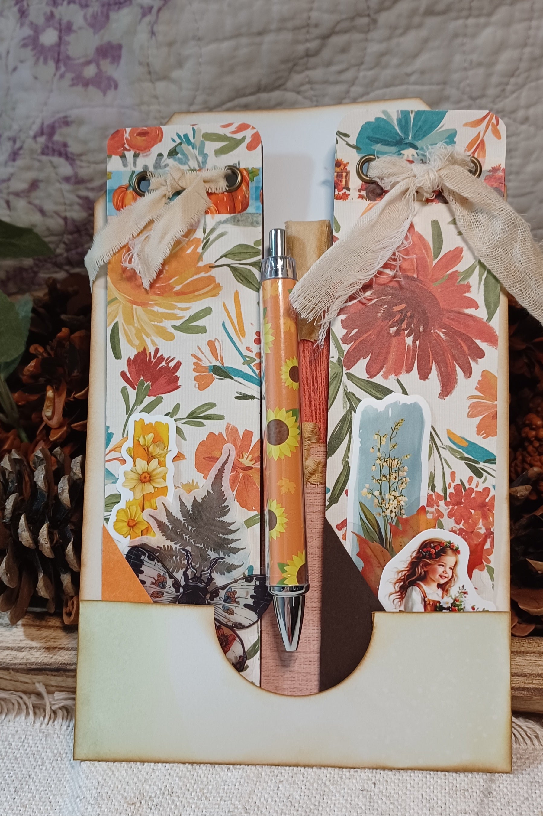 A decorative gift set featuring a floral and rustic-themed card holder with two matching floral cards tied with beige ribbon, holding a sunflower-patterned pen and a wooden stick, displayed on a textured fabric surface with pinecones in the background.