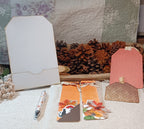 A styled flat-lay of handmade paper craft items, including two orange autumn-themed bookmark cards with ribbons, a blank white cardstock tag, a pink textured tag, a brown lace tag, and a patterned pen, arranged on a white textured blanket with pinecones and a quilt in the background.