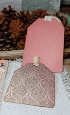 Two decorative wooden gift tags with a small loop at the top, displayed on a textured fabric with pinecones in the background. One tag is a solid dusty pink, and the other features an embossed floral pattern in a muted mauve-brown tone.