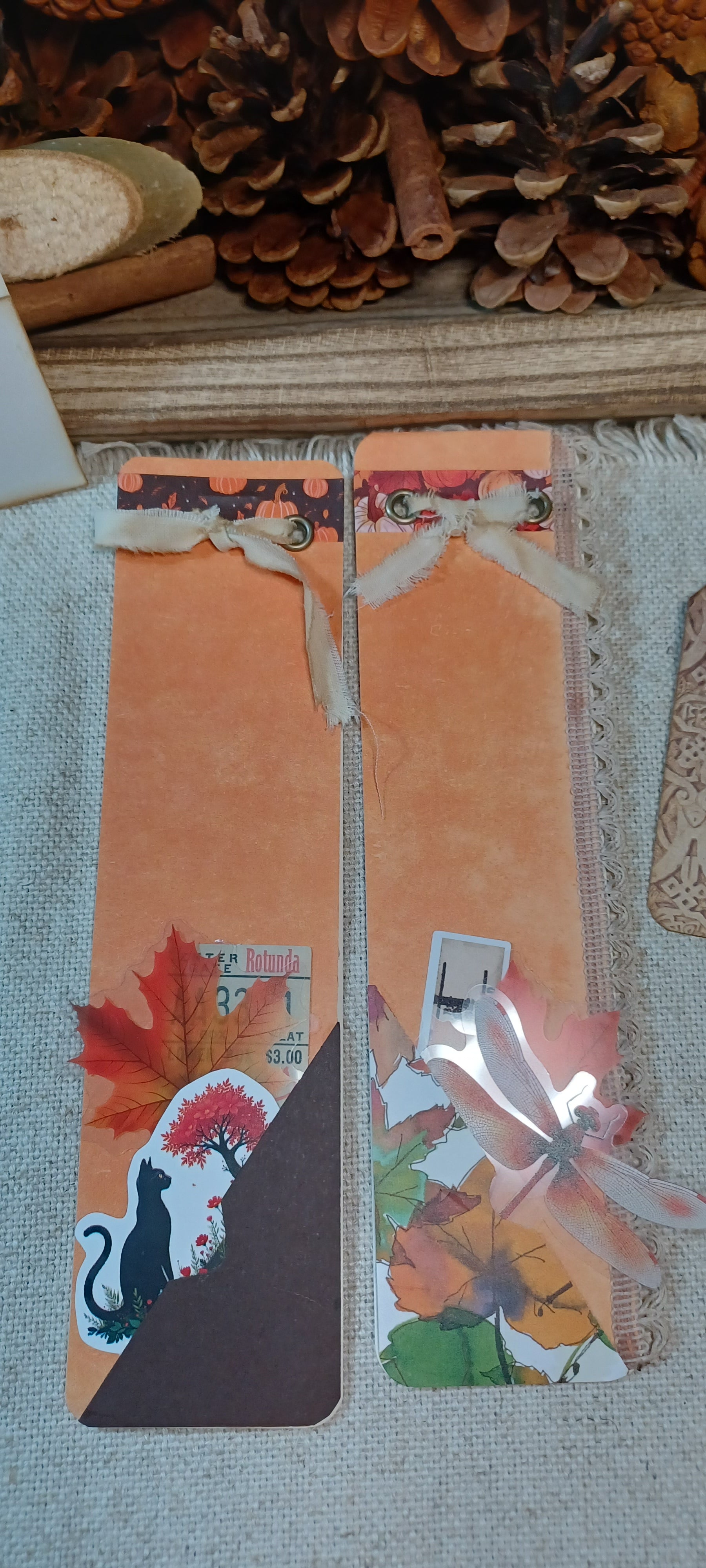 Two handmade autumn-themed bookmark cards displayed on a textured fabric surface with pinecones and wood in the background. The bookmarks feature orange backgrounds, colorful fall leaves, a black cat silhouette, and ribbon bows.
