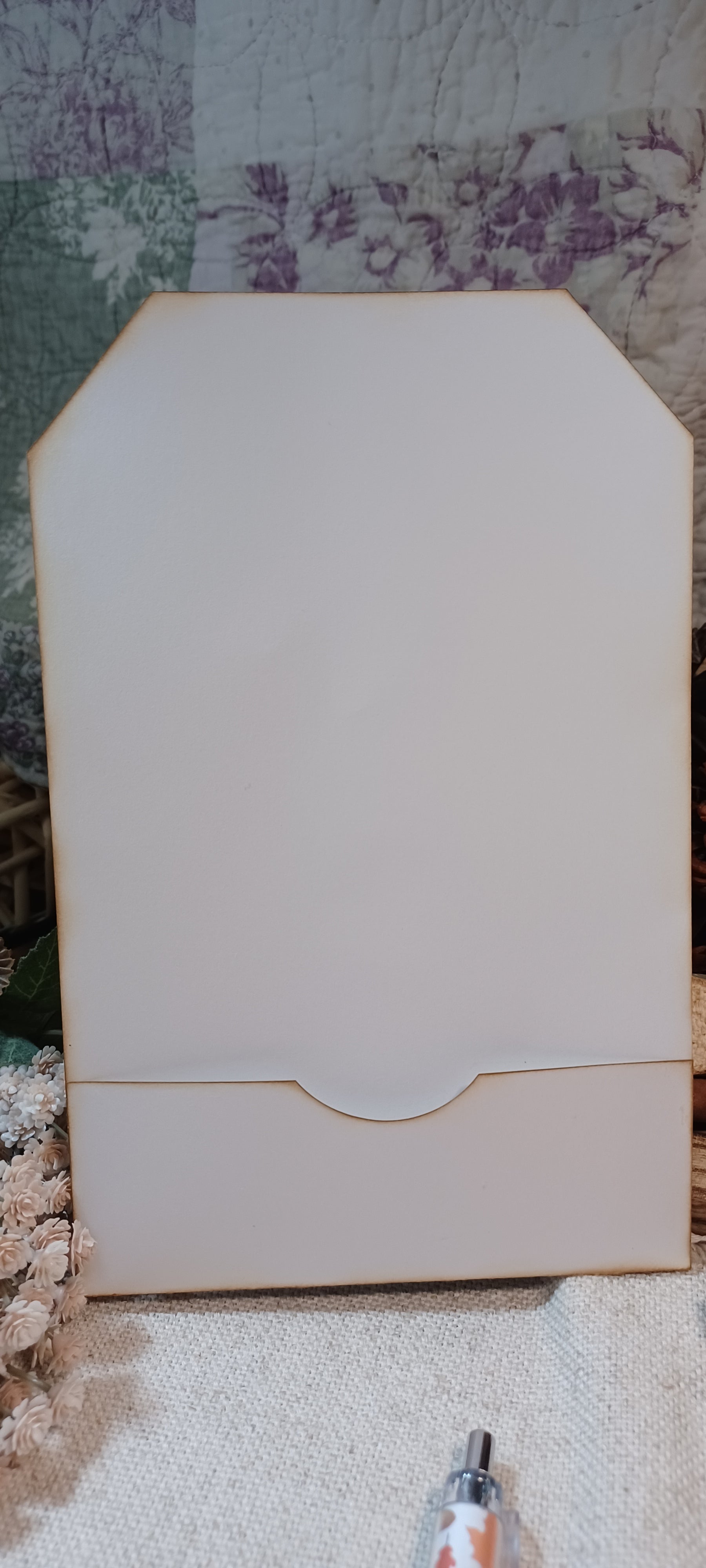 A large, blank, off-white wooden display card or sign with a small drawer cutout at the bottom, standing upright on a textured fabric surface.