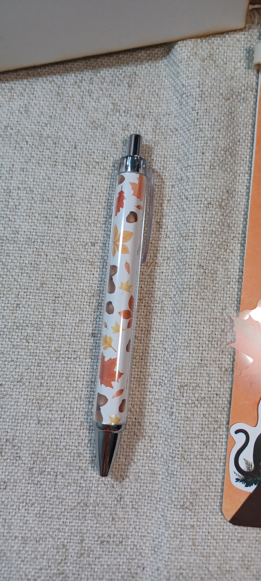A clear pen with a white background and an autumn leaf pattern in orange, brown, and yellow, resting on a textured beige fabric surface.