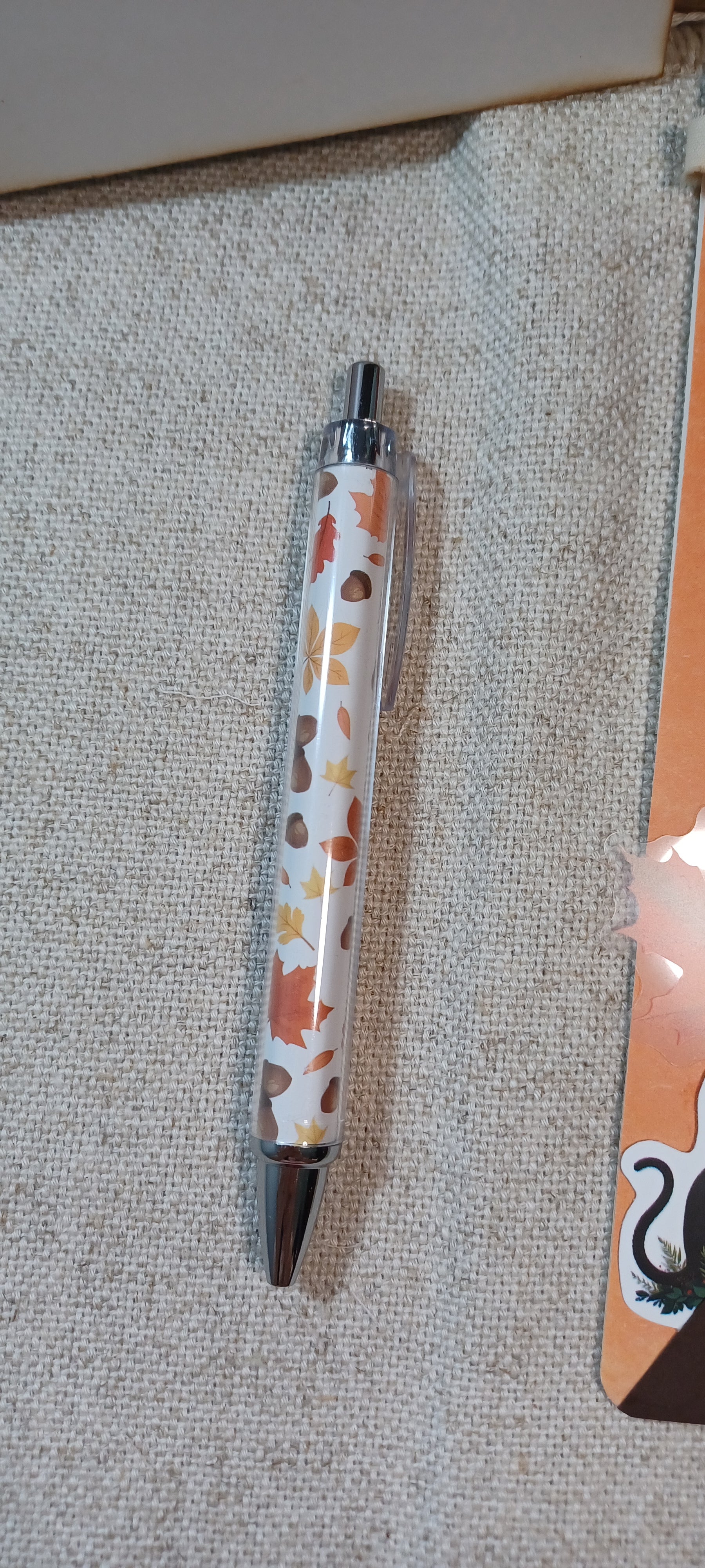 A clear pen with a white background and an autumn leaf pattern in orange, brown, and yellow, resting on a textured beige fabric surface.