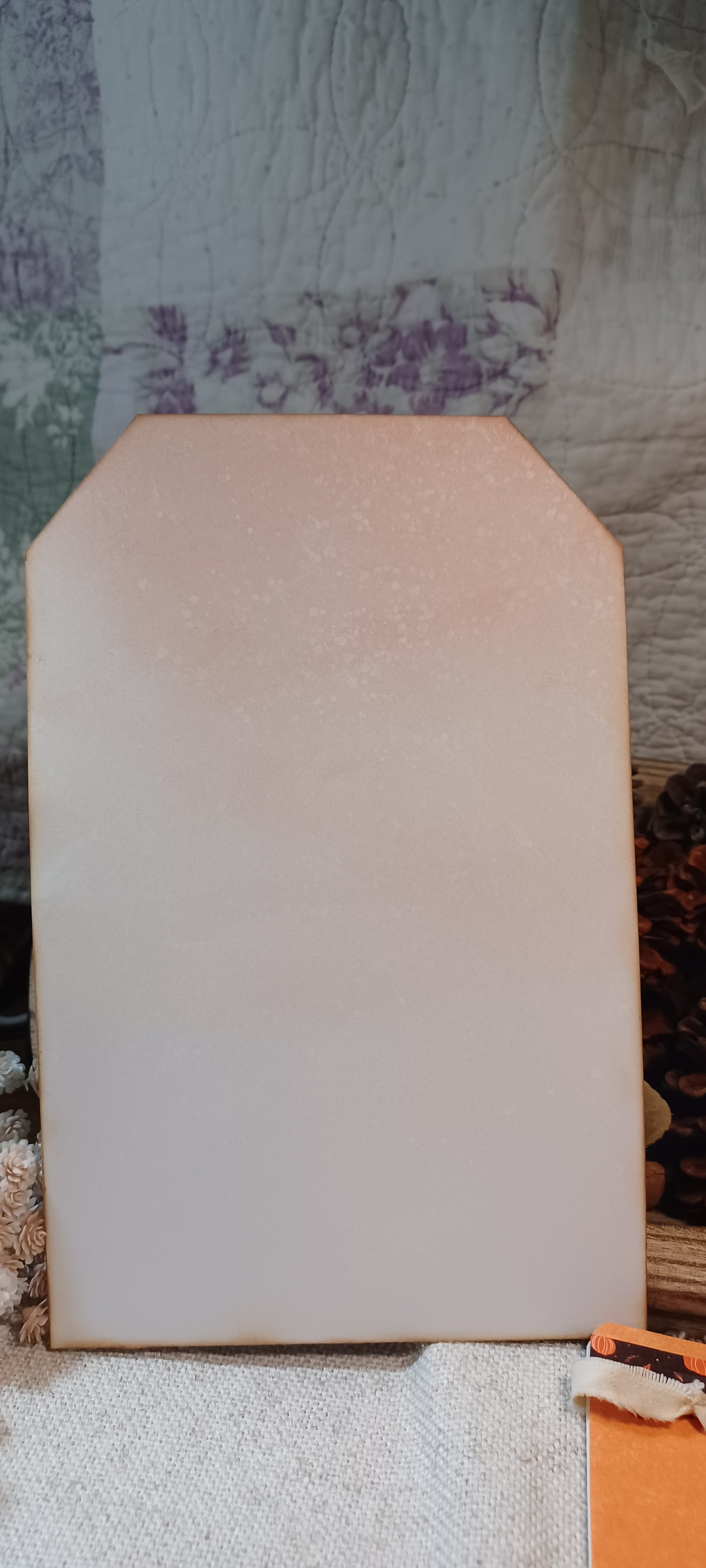 A large, rectangular, light pink decorative board or sign with a scalloped top edge, standing upright on a textured surface against a patterned fabric background.