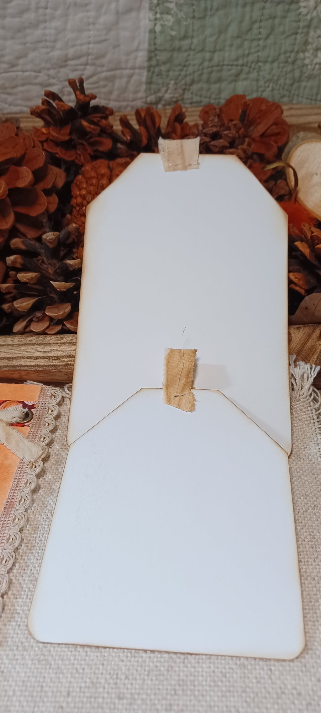 A blank white wooden sign or board with a small piece of tape at the top and center, displayed on a textured fabric surface with pinecones and autumn decorations in the background.