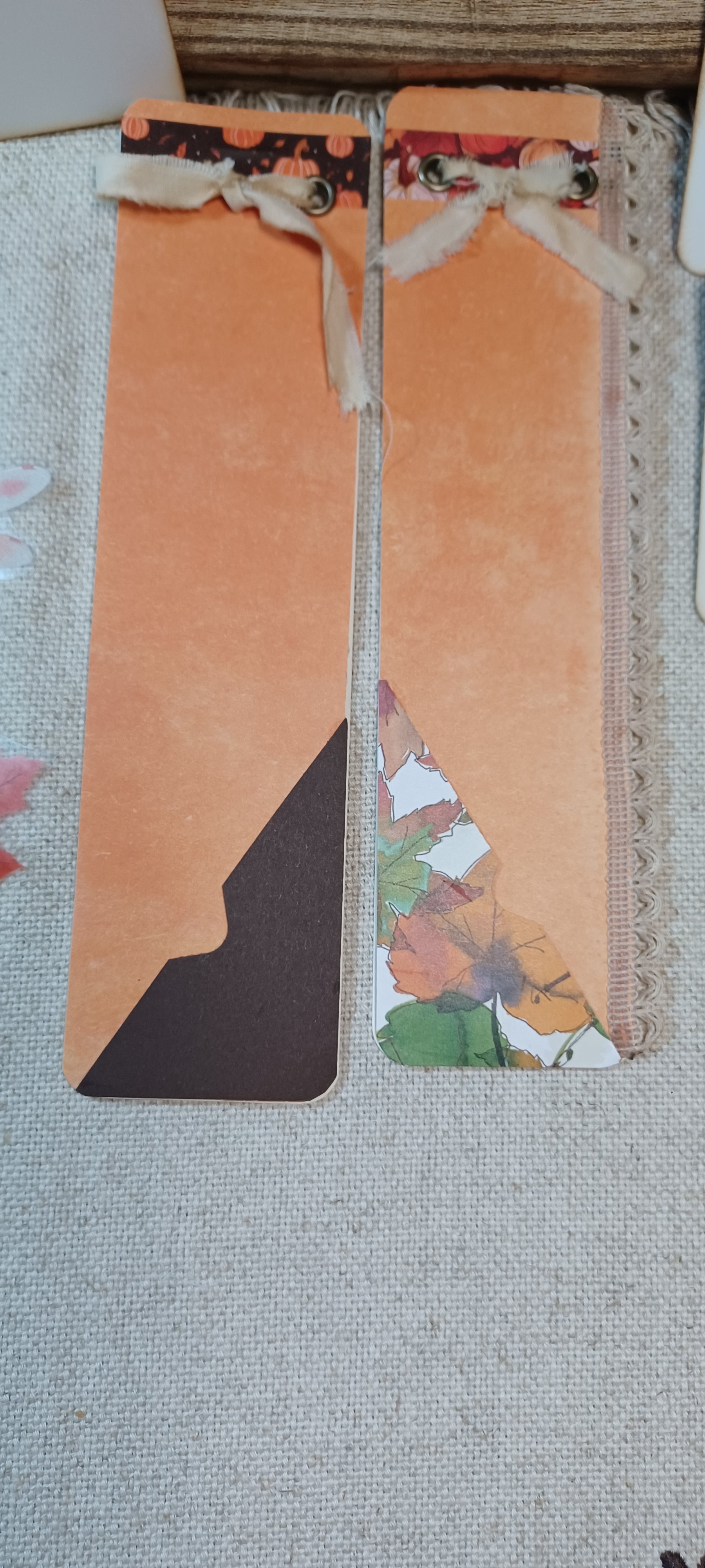 Two decorative paper bookmarks with orange panels, floral patterned accents, and ribbon bows, laid on a light fabric surface.