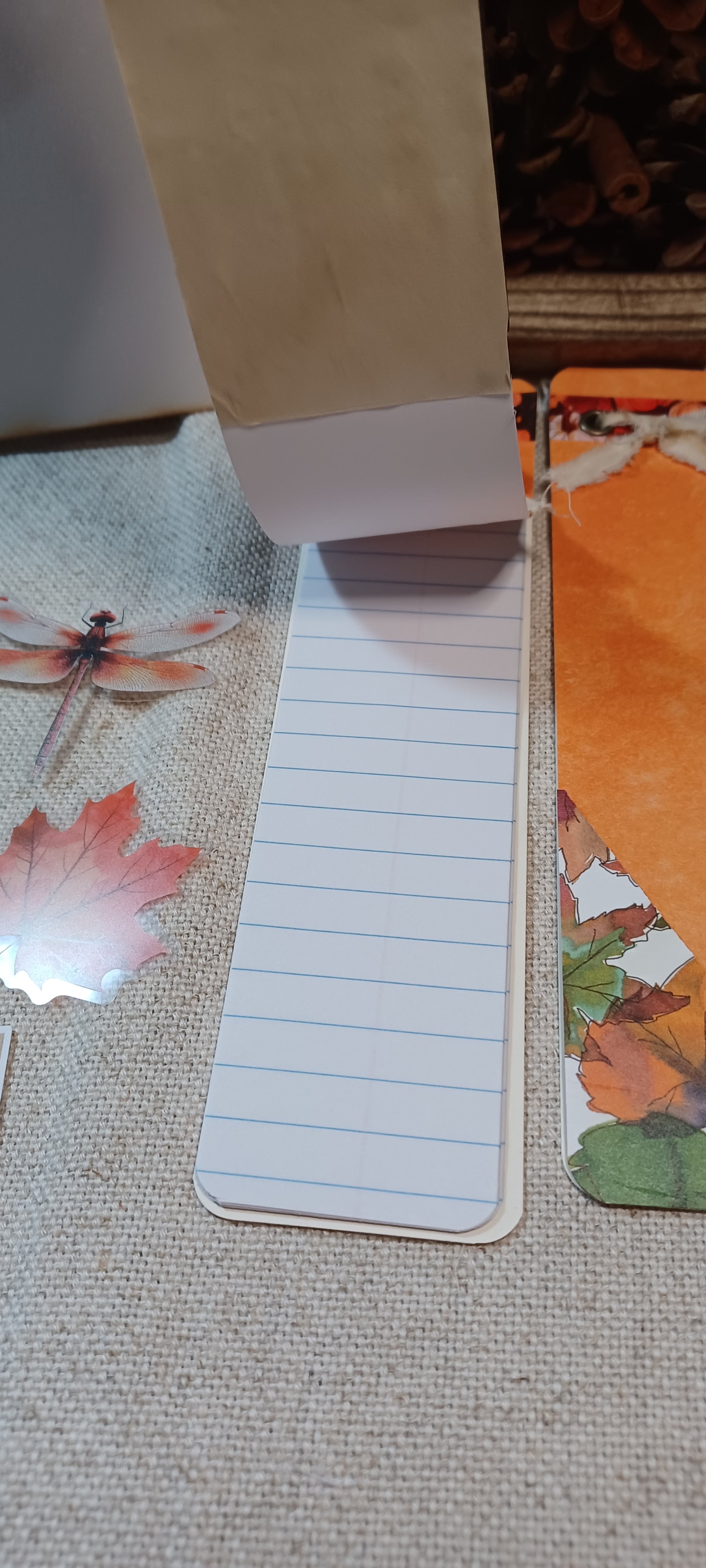 A close-up of a lined notepad or notebook page with a brown paper pocket attached at the top, placed on a textured fabric surface with autumn leaf decorations.