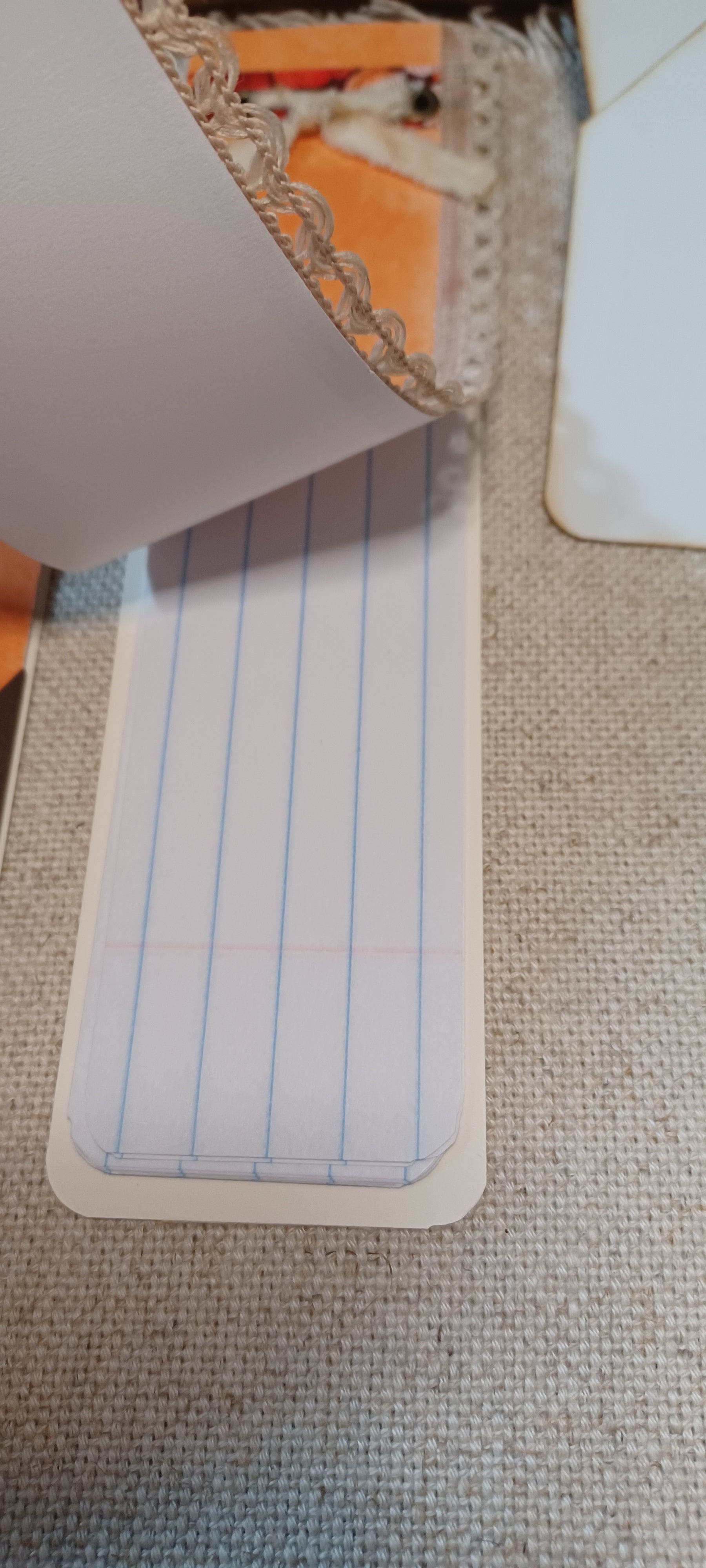 A close-up of a lined notepad page with blue vertical lines and a beige border, partially covered by a white sheet, placed on a textured beige fabric surface.