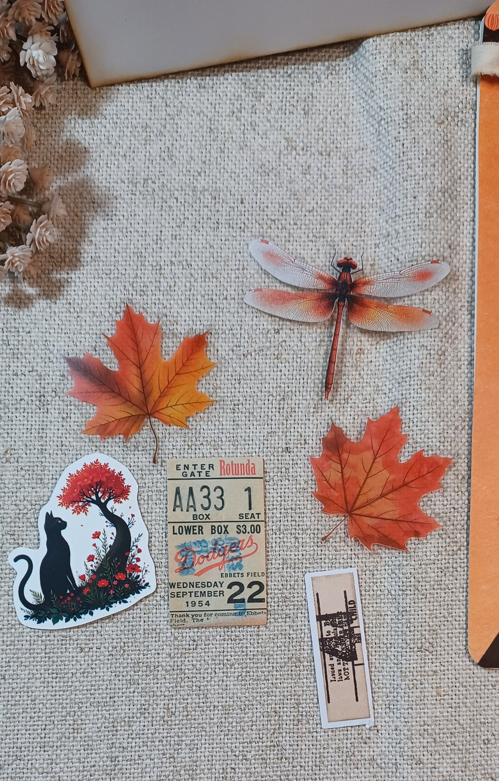A collection of autumn-themed stickers arranged on a beige burlap fabric background, featuring orange maple leaves, a dragonfly, a black cat under a tree, a vintage ticket, and a small architectural ticket.