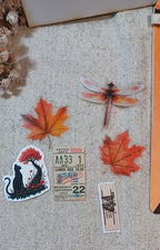 A collection of autumn-themed stickers arranged on a beige burlap fabric background, featuring orange maple leaves, a dragonfly, a black cat under a tree, a vintage ticket, and a small architectural ticket.