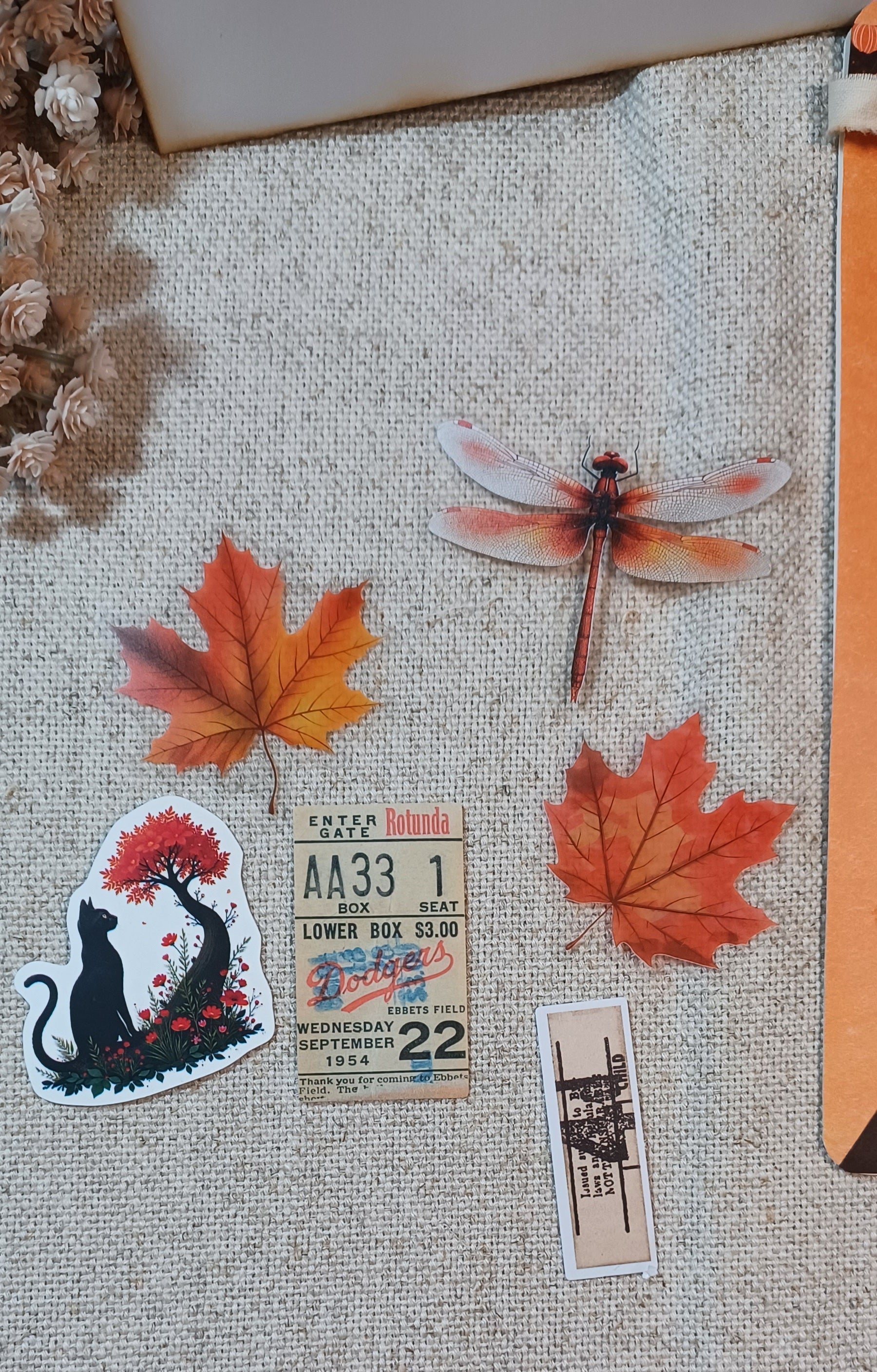 A collection of autumn-themed stickers arranged on a beige burlap fabric background, featuring orange maple leaves, a dragonfly, a black cat under a tree, a vintage ticket, and a small architectural ticket.