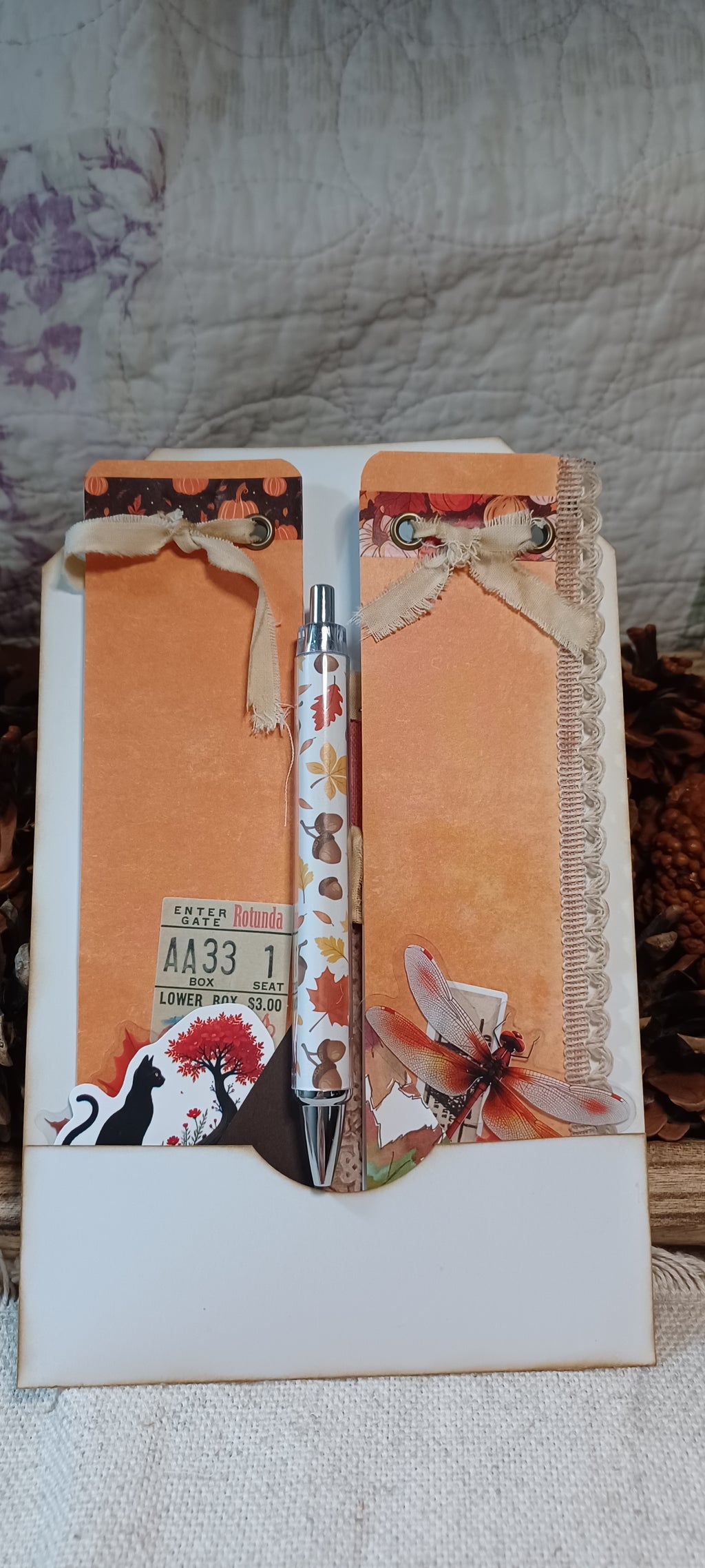 A decorative pen set inside a white envelope-style holder with autumn-themed embellishments, including orange textured paper, lace ribbon, a small ticket sticker, and a butterfly illustration, photographed on a fabric surface.