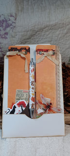 A decorative pen set inside a white envelope-style holder with autumn-themed embellishments, including orange textured paper, lace ribbon, a small ticket sticker, and a butterfly illustration, photographed on a fabric surface.