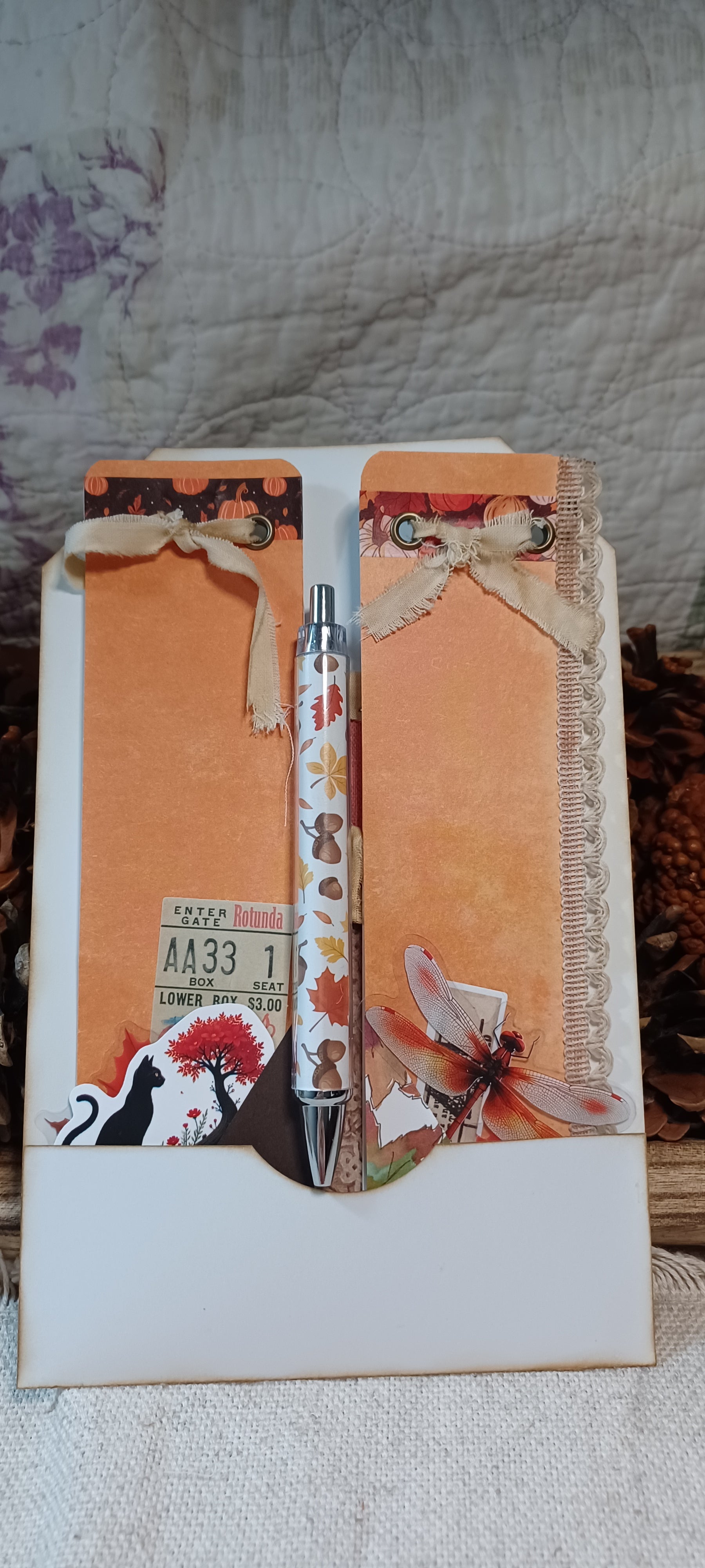 A decorative pen set inside a white envelope-style holder with autumn-themed embellishments, including orange textured paper, lace ribbon, a small ticket sticker, and a butterfly illustration, photographed on a fabric surface.