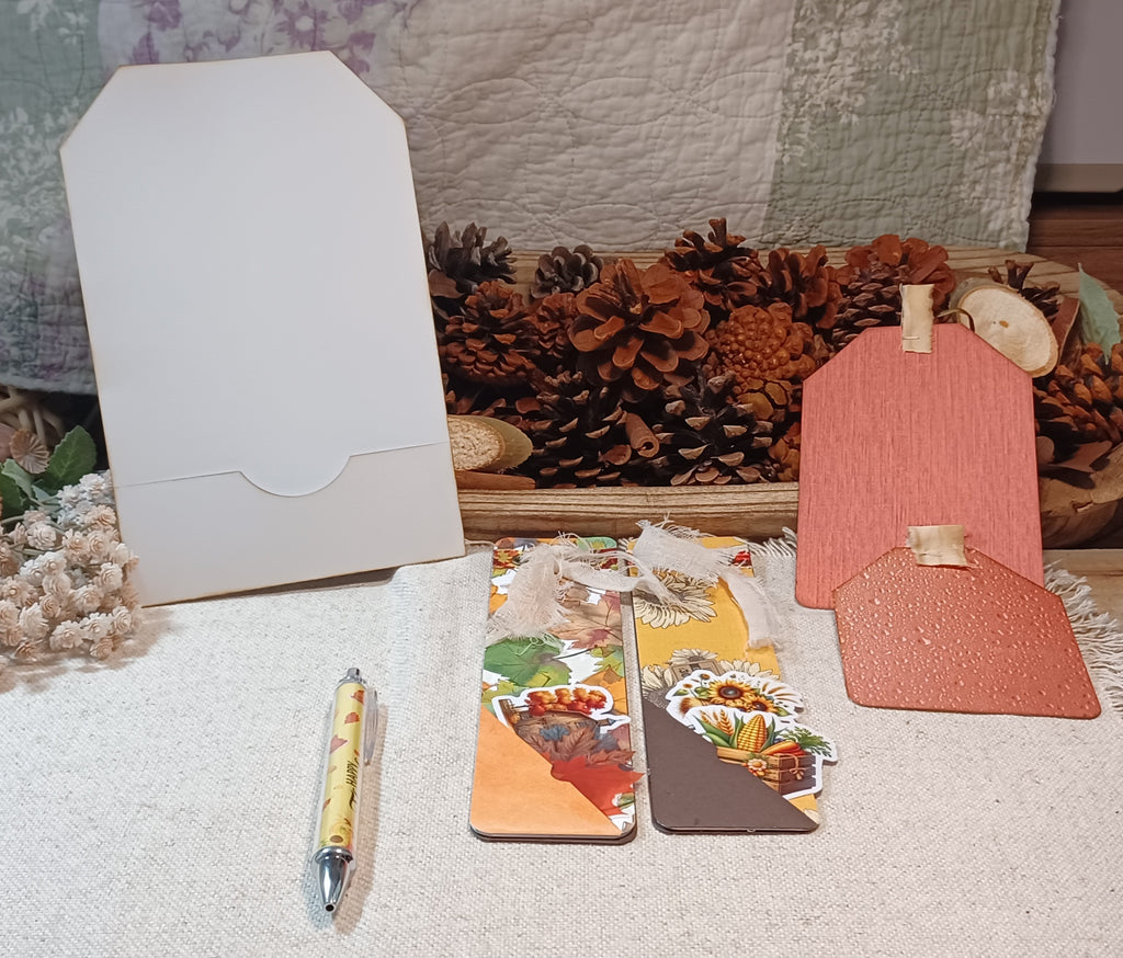 A styled flat-lay of handmade paper goods, including a white envelope, two decorative paper bookmarks with autumn-themed illustrations, a pink textured paper tag, and a yellow patterned pen, arranged on a white textured fabric with pine cones and a quilted blanket in the background.
