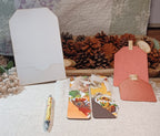 A styled flat-lay of handmade paper goods, including a white envelope, two decorative paper bookmarks with autumn-themed illustrations, a pink textured paper tag, and a yellow patterned pen, arranged on a white textured fabric with pine cones and a quilted blanket in the background.