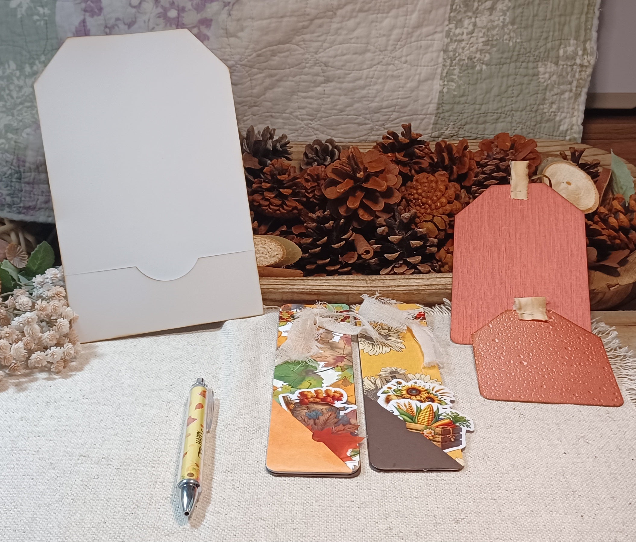 A styled flat-lay of handmade paper goods, including a white envelope, two decorative paper bookmarks with autumn-themed illustrations, a pink textured paper tag, and a yellow patterned pen, arranged on a white textured fabric with pine cones and a quilted blanket in the background.