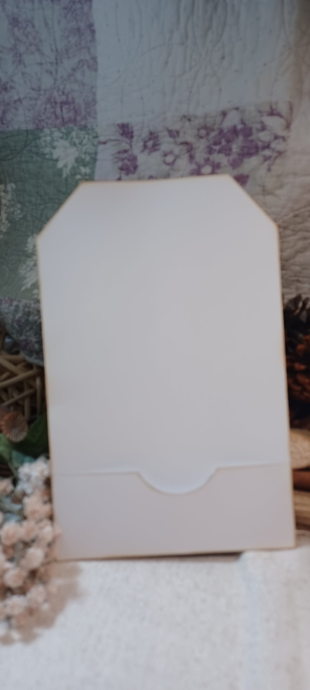 A blank, unbranded white envelope or card holder with a small tab closure at the top, standing upright on a textured white surface against a floral patchwork fabric background.