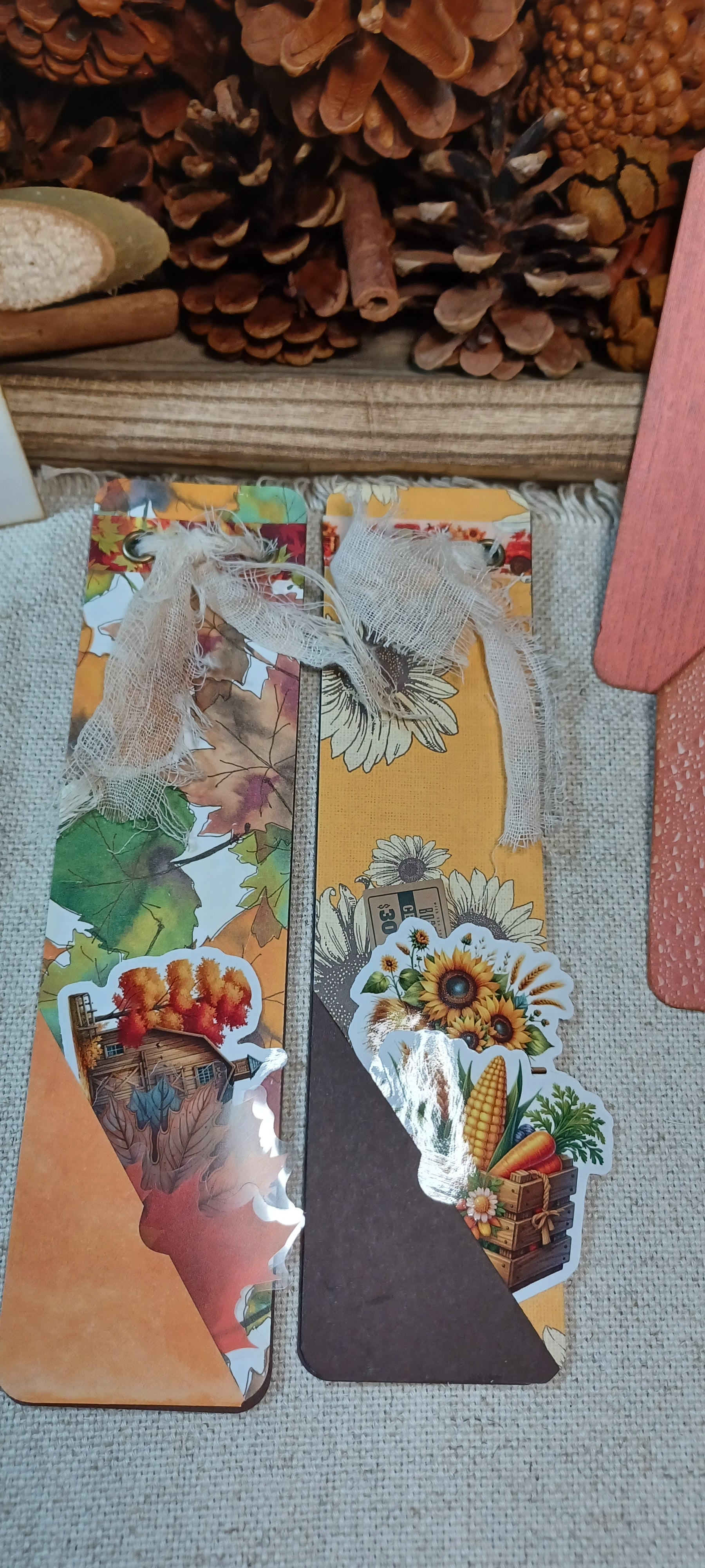 A handmade autumn-themed paper craft or scrapbook card featuring layered collage elements including sunflowers, pinecones, leaves, and rustic fall illustrations, displayed on a neutral fabric surface with pinecones in the background.