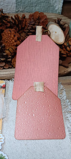Two dusty pink textured paper gift tags with small beige ribbon loops, displayed on a neutral fabric surface with pinecones and wood decor in the background.