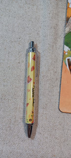 A yellow retractable pen with a leaf pattern and the word 'HAPPY' printed vertically, lying on a textured beige fabric surface.