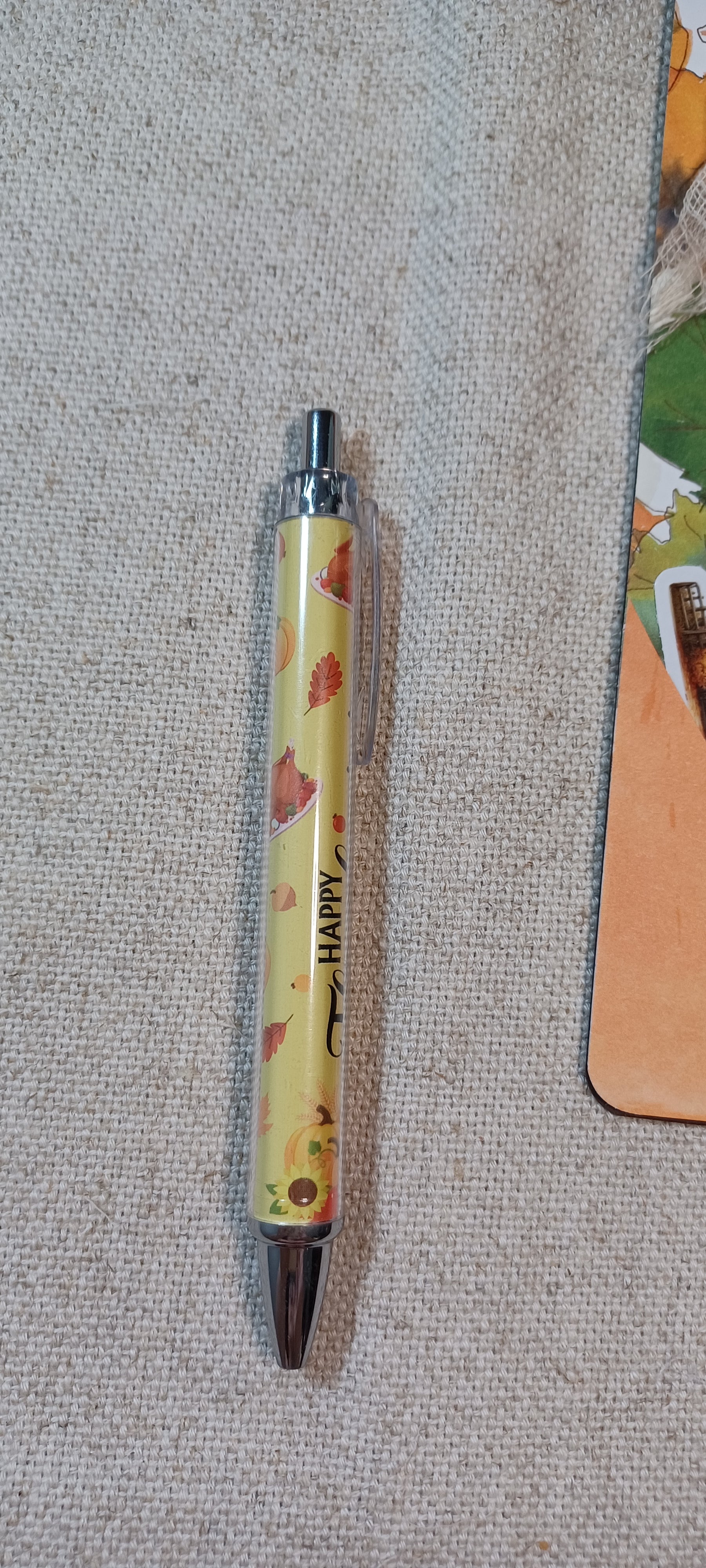 A yellow retractable pen with a leaf pattern and the word 'HAPPY' printed vertically, lying on a textured beige fabric surface.