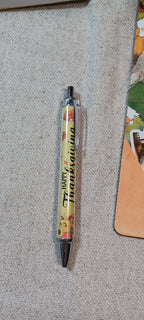 A pen with a Thanksgiving-themed design, featuring a yellow body with autumn leaves and the text 'Happy Thanksgiving' in black script, resting on a textured beige fabric surface.
