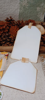 Two blank white wooden sign boards with natural wood edges and small wooden tabs, displayed on a burlap surface with pinecones and a quilted fabric backdrop.