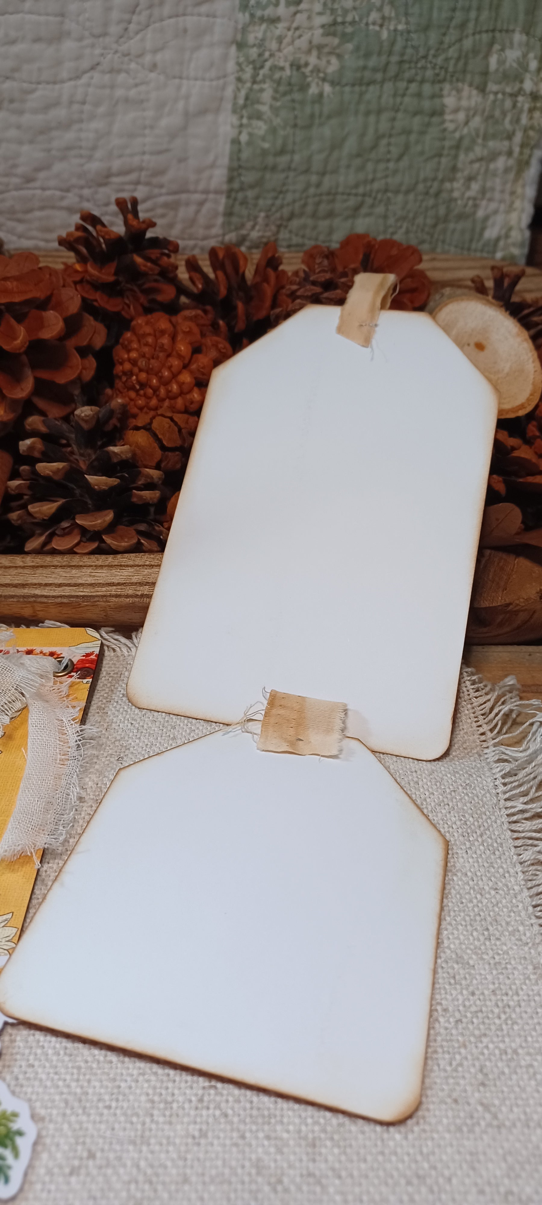 Two blank white wooden sign boards with natural wood edges and small wooden tabs, displayed on a burlap surface with pinecones and a quilted fabric backdrop.