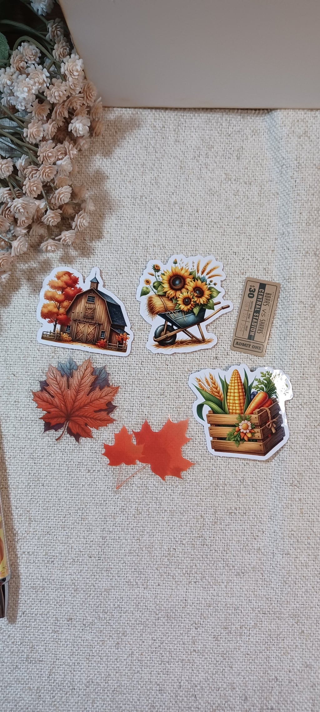 Five autumn-themed stickers are arranged on a beige textured fabric surface. The stickers depict a barn with fall foliage, sunflowers in a wheelbarrow, red maple leaves, and a corn basket.