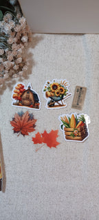 Five autumn-themed stickers are arranged on a beige textured fabric surface. The stickers depict a barn with fall foliage, sunflowers in a wheelbarrow, red maple leaves, and a corn basket.