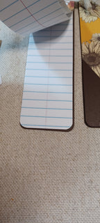A close-up of a lined notepad sheet with blue horizontal lines and rounded corners, placed on a beige woven fabric surface next to other paper items.
